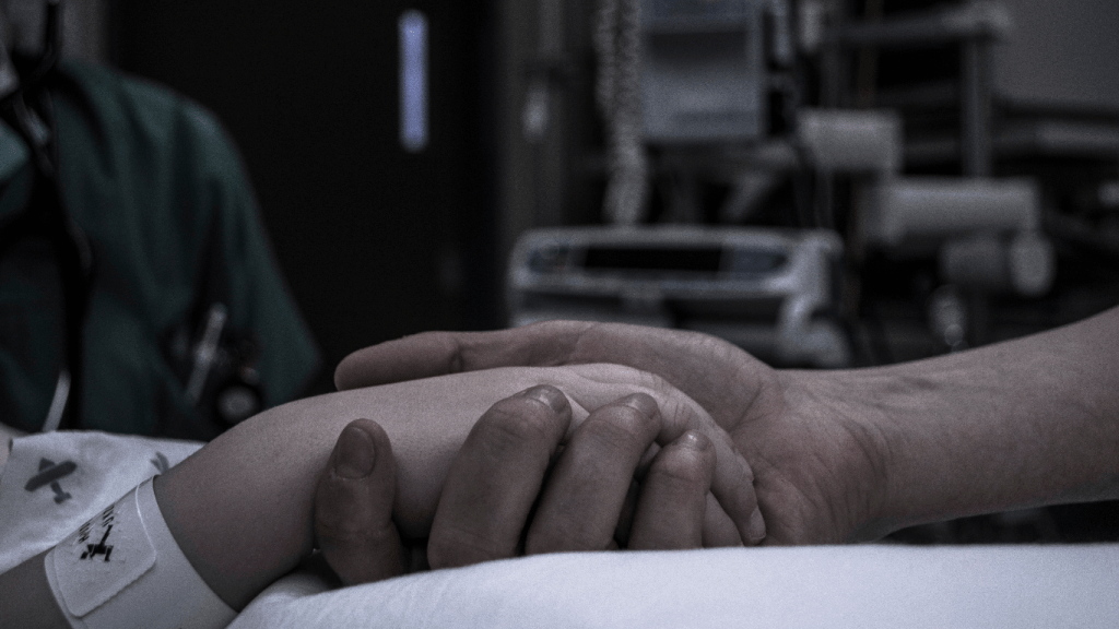 Man holding his dying wife's hand in a hospital bed while she is lying unconscious 
